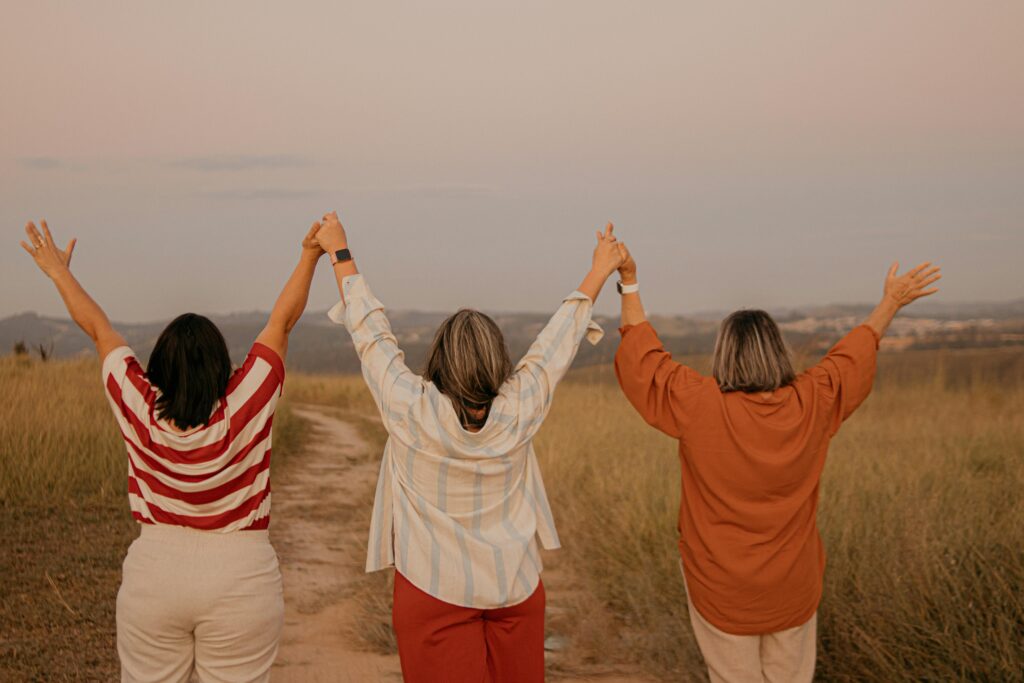 Three women standing in a São Paulo field, holding hands and raising arms, celebrating friendship.