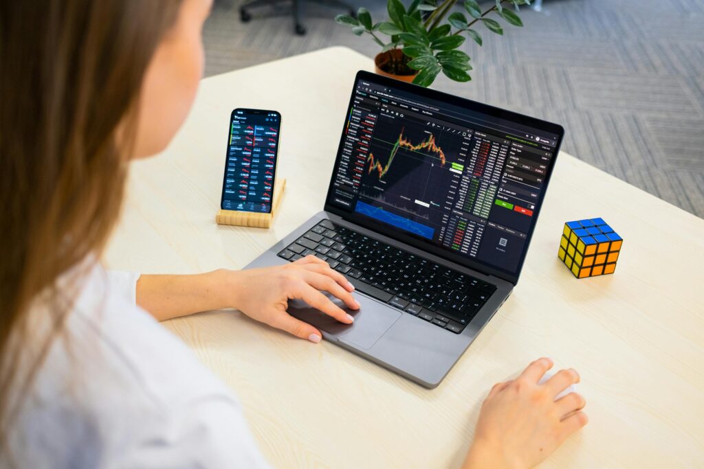 How To Start Trading Crypto: A Beginners Checklist A woman using a laptop for stock trading analysis with a smartphone and Rubik's Cube nearby.
