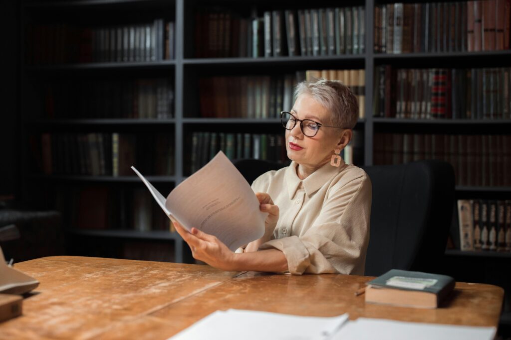 What Every Crypto Mama Needs to Know Before She Buys Her First Bitcoin Elegant senior woman reading a manuscript at a wooden desk in a cozy library setting, surrounded by bookshelves.