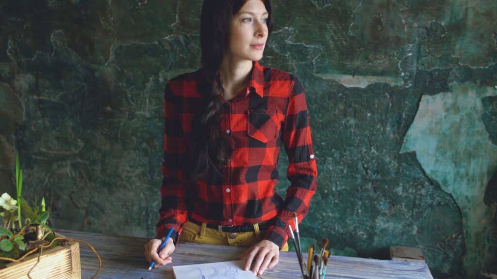 Woman in red plaid shirt sketching at a wooden table, exuding creativity.