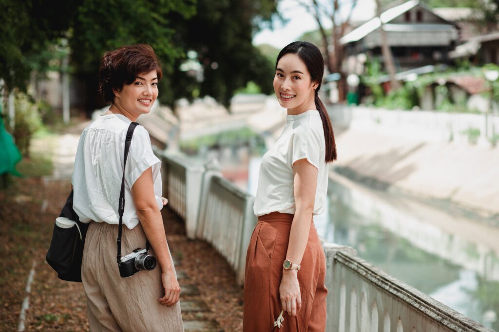 Back view of young cheerful ethnic female friends in casual wear looking at camera over shoulder near water channel in summer