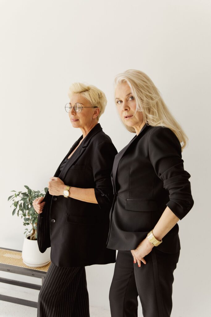 Two fashionable mature women in black blazers posing inside an elegant setting, exuding style and confidence.